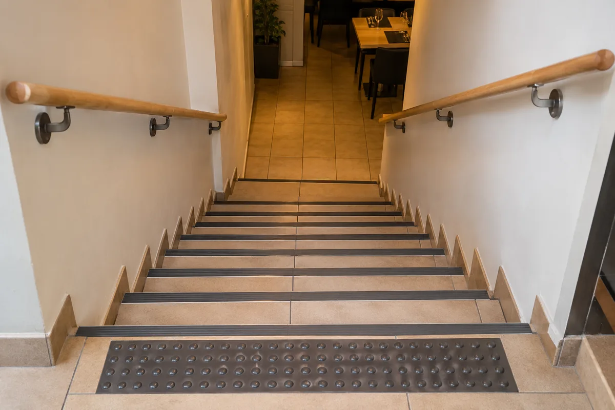 Escalier carrelé de restaurant descendant vers le sous-sol, nez de marche en aluminium contrastés et main courante en bois